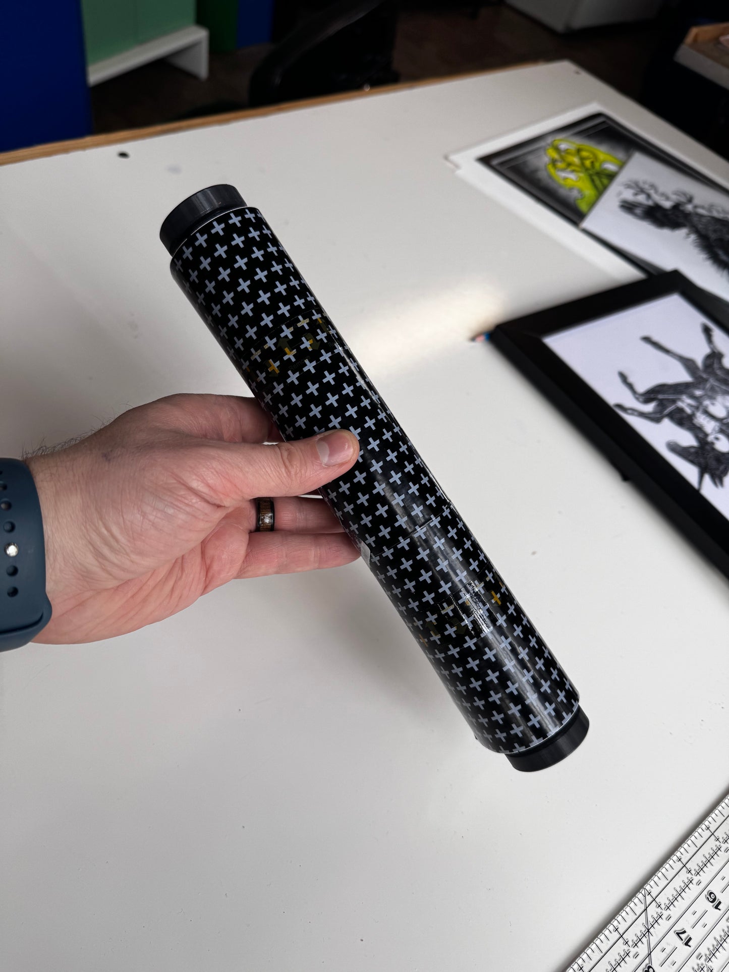 Hand holding a roll of tattoo bandage above a white surface with framed pictures in the background.