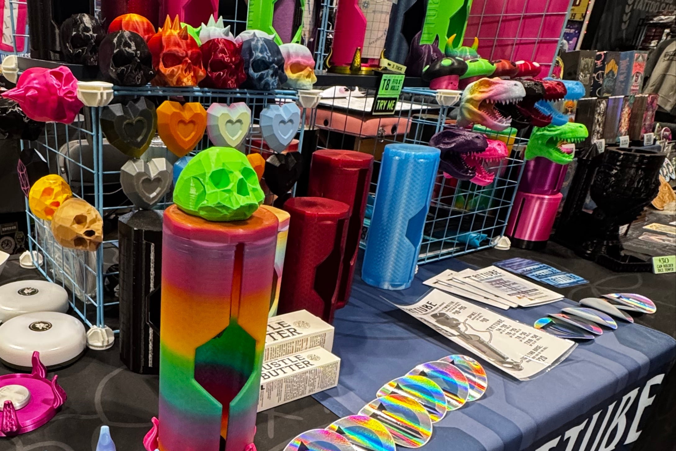 Colorful 3D-printed skulls, dinosaur heads, mushrooms, and geometric hearts. 3D-printed cylinders for dispensing tattoo bandages in various colors and sizes. Stickers and information on a trade show table. 
