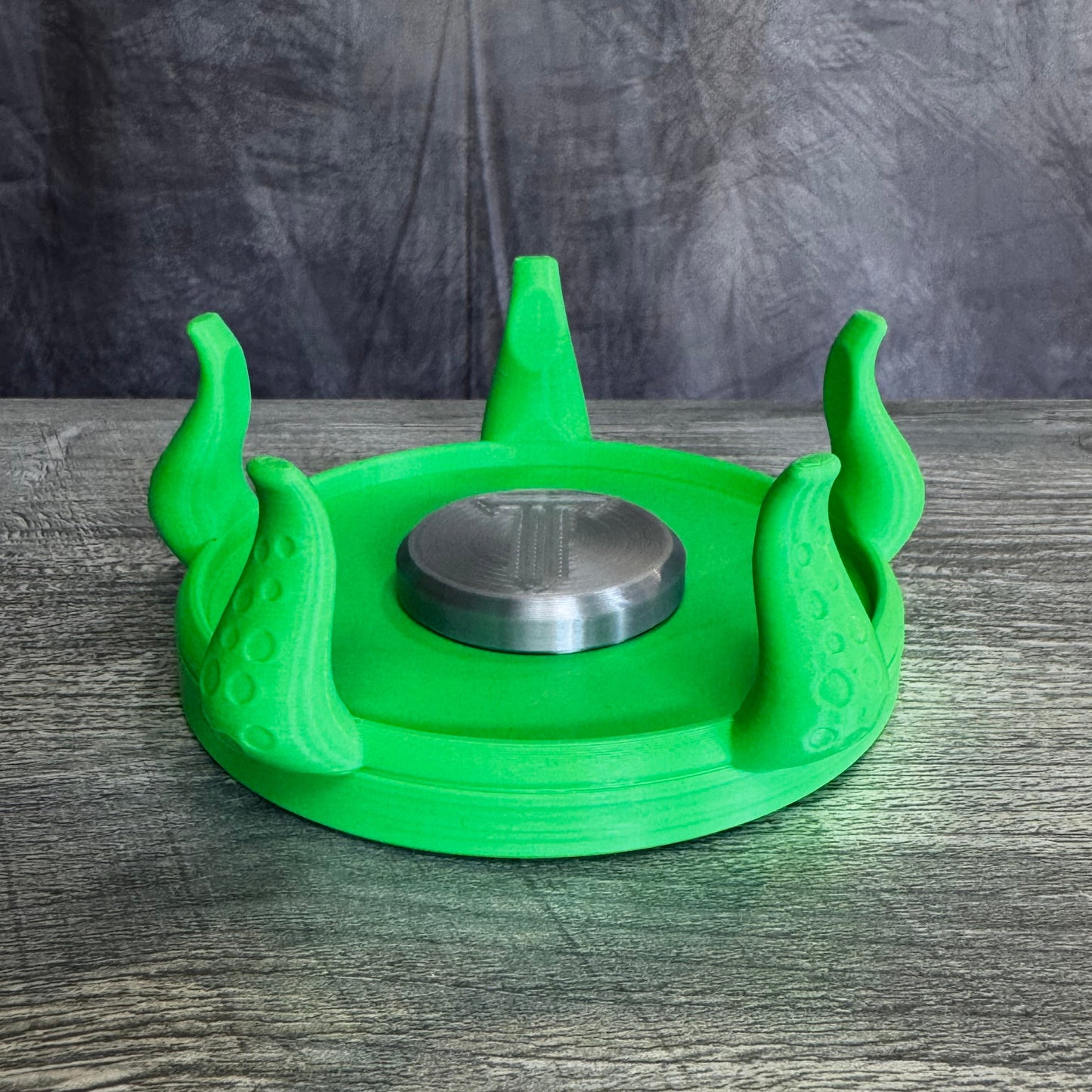 Black plastic base with five tentacles, with silver plastic disc on the middle, sitting gray wood table.